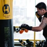 Techniques - Full length side view of barefoot Asian boxer in gloves punching bag during workout in light gym