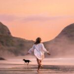 Lengths - Back view full length slim female in white males shirt running with dog on scenic wet seacoast against rough cliffs on early evening