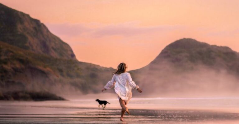 Lengths - Back view full length slim female in white males shirt running with dog on scenic wet seacoast against rough cliffs on early evening