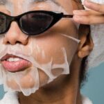 Routine - Self esteem young African American lady with moisturizing sheet mask and towel on hand wearing trendy sunglasses while standing against blue background in bathrobe after shower