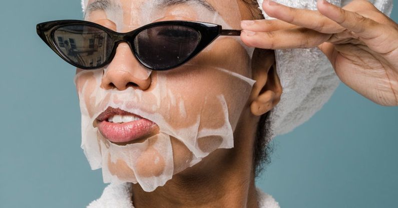 Routine - Self esteem young African American lady with moisturizing sheet mask and towel on hand wearing trendy sunglasses while standing against blue background in bathrobe after shower