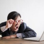 Mistakes - Modern Asian man in jacket and glasses looking at laptop and screaming with mouth wide opened on white background