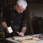 Techniques - Serious male woodworker in apron using mallet and chisel to carve wooden board at table with abundance of instruments in carpentry