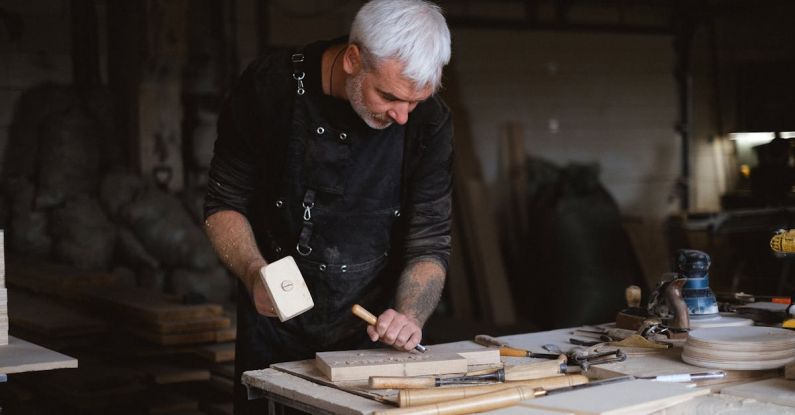 Techniques - Serious male woodworker in apron using mallet and chisel to carve wooden board at table with abundance of instruments in carpentry