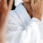 Routine - Young black female in white robe and towel on head applying moisturizing cream on face while standing in bathroom