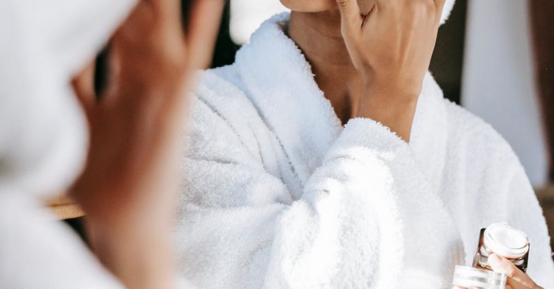 Routine - Young black female in white robe and towel on head applying moisturizing cream on face while standing in bathroom